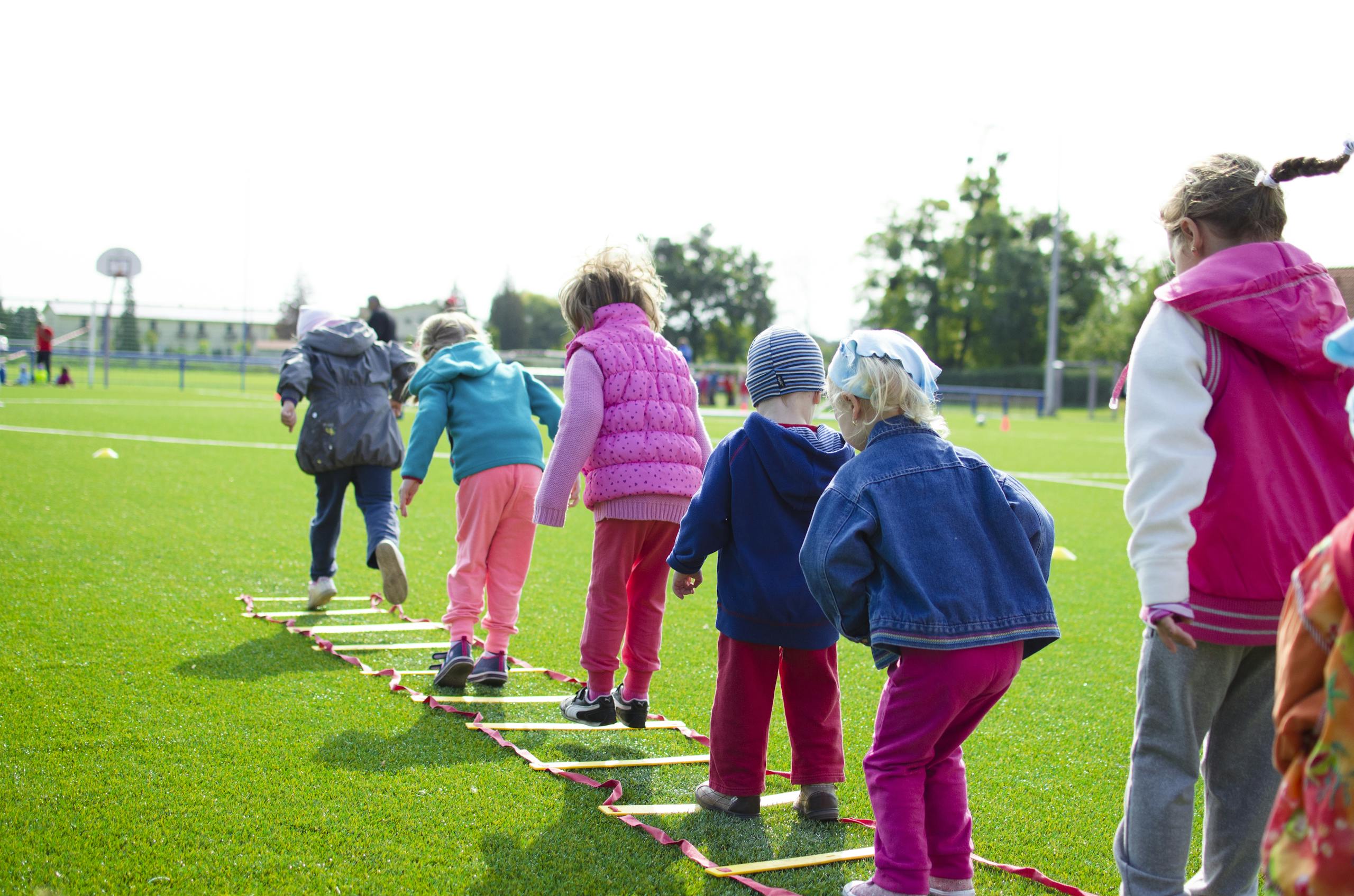 Children enjoy an outdoor activity on a grassy field, stepping over a ladder.